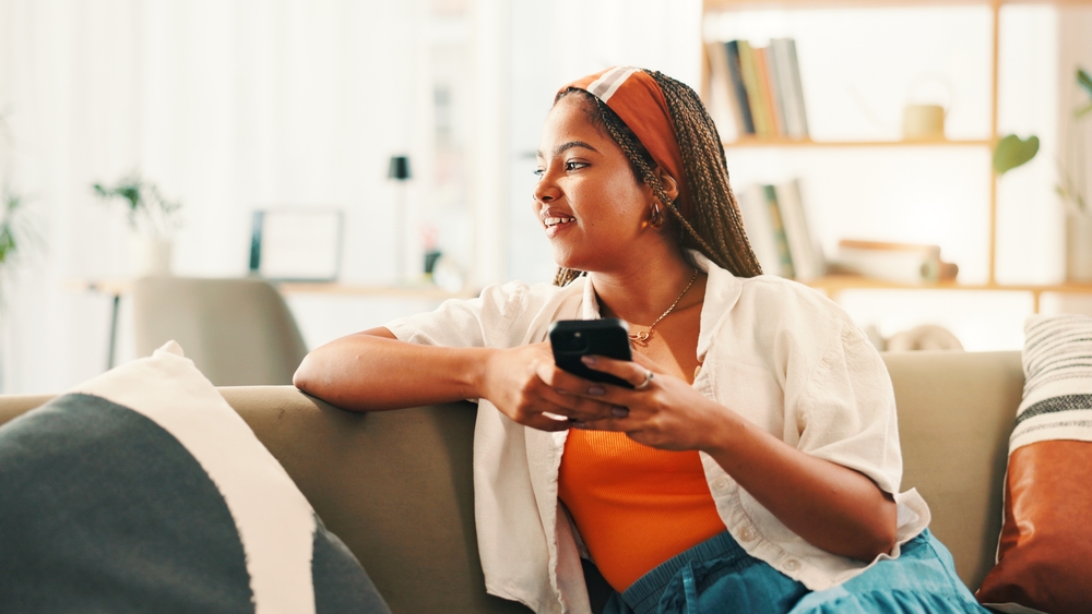 A woman with braided hair sits on the couch, holding her mobile phone.