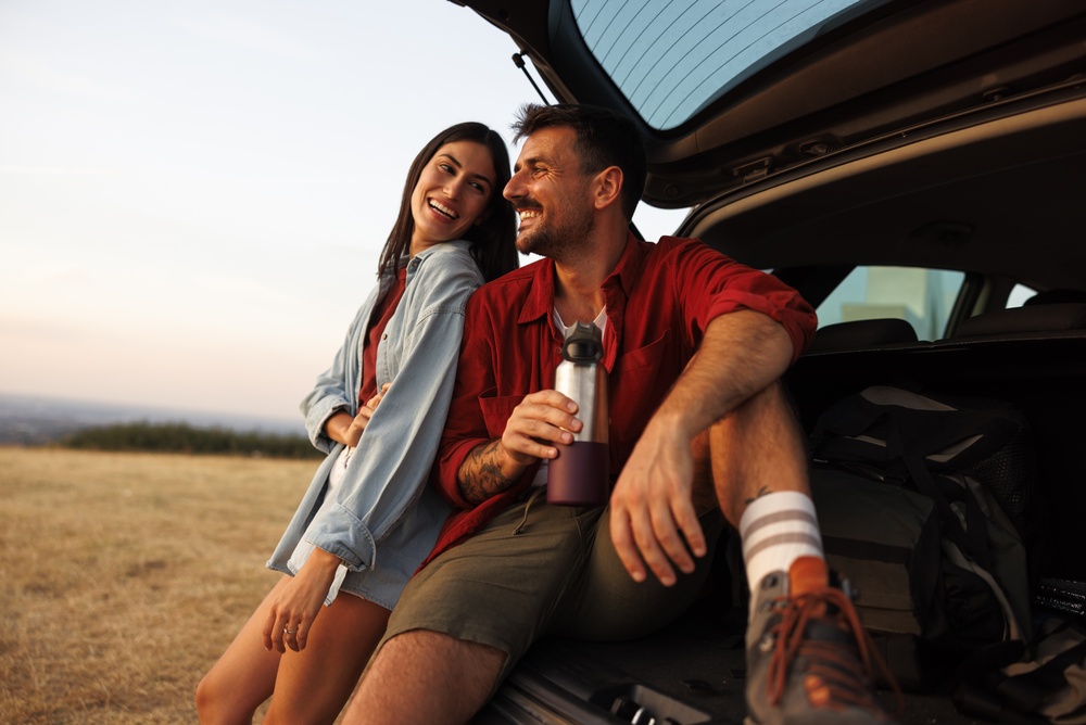 A man and a woman in hiking clothes relax on the tailgate of their SUV after a long road trip.