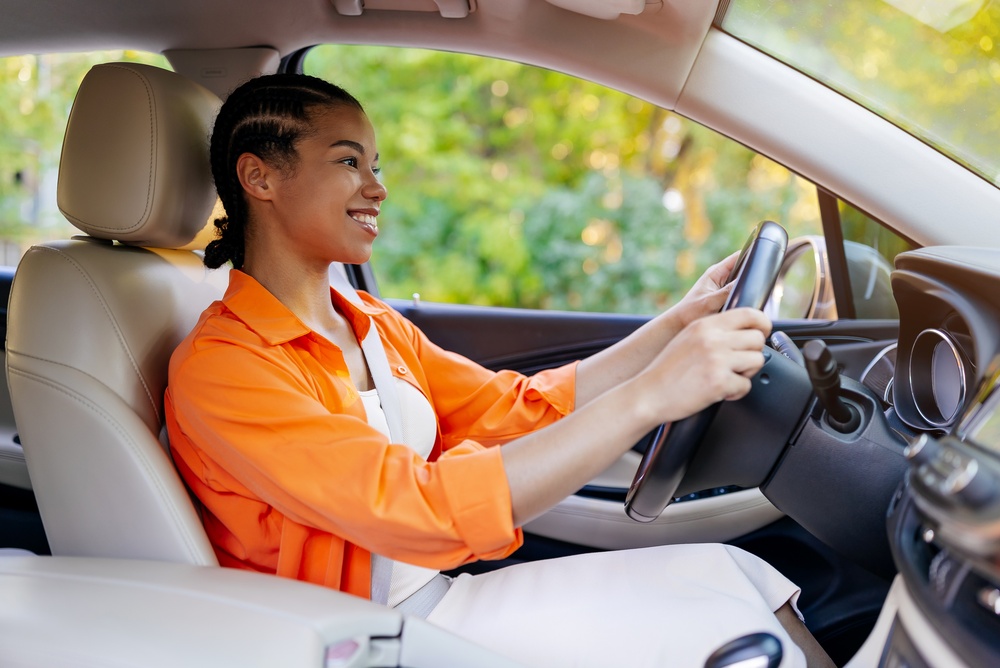 A smiling young woman wearing an orange blouse sits behind the wheel of her car.