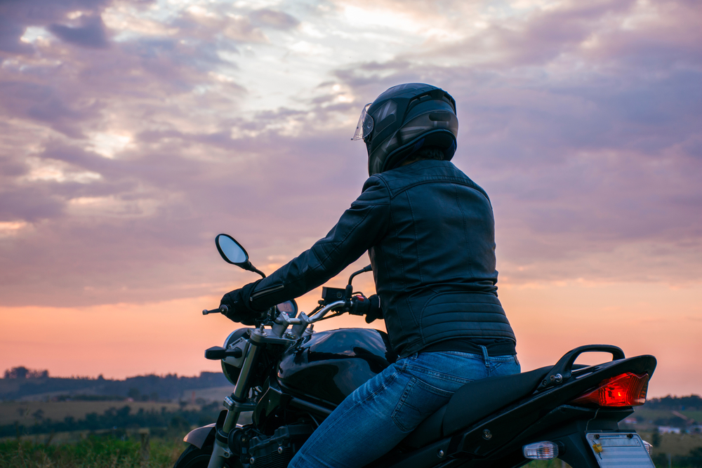 A man in a helmet and black leather coat sits on a motorcycle at sunset.