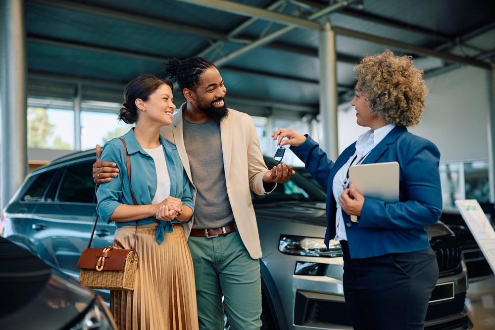 A man and a woman smile as they accept the keys to their new car from the dealership sales representative.