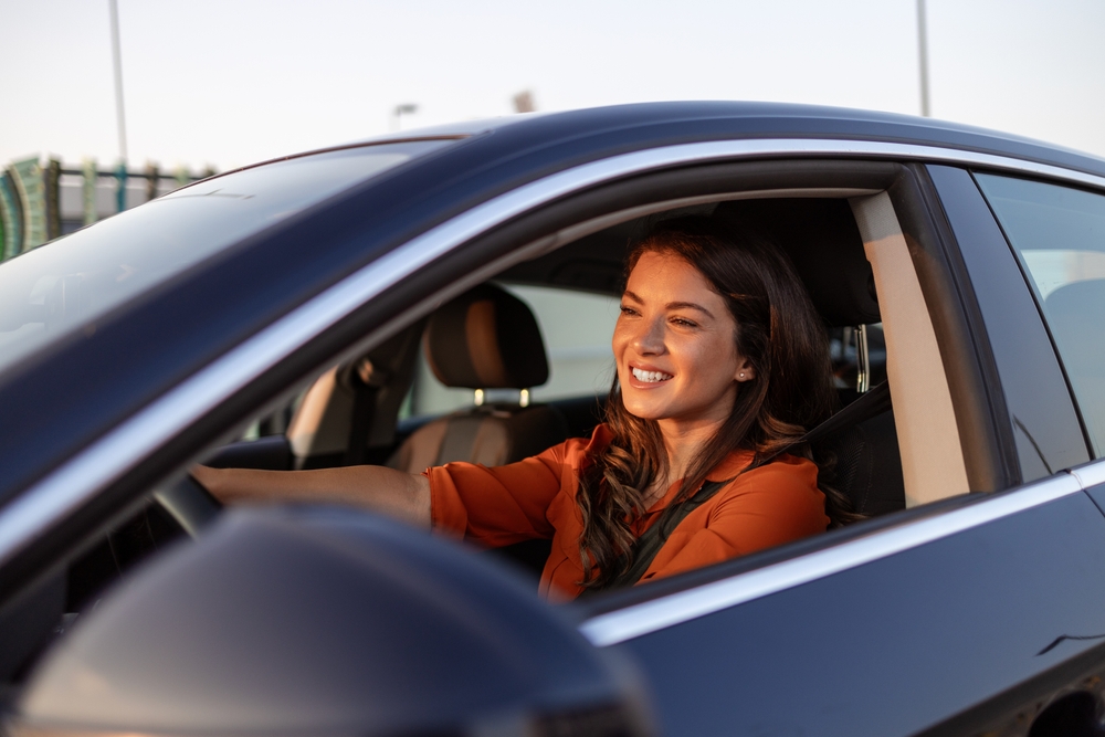A woman with brown hair smiles as she drives a dark blue car.
