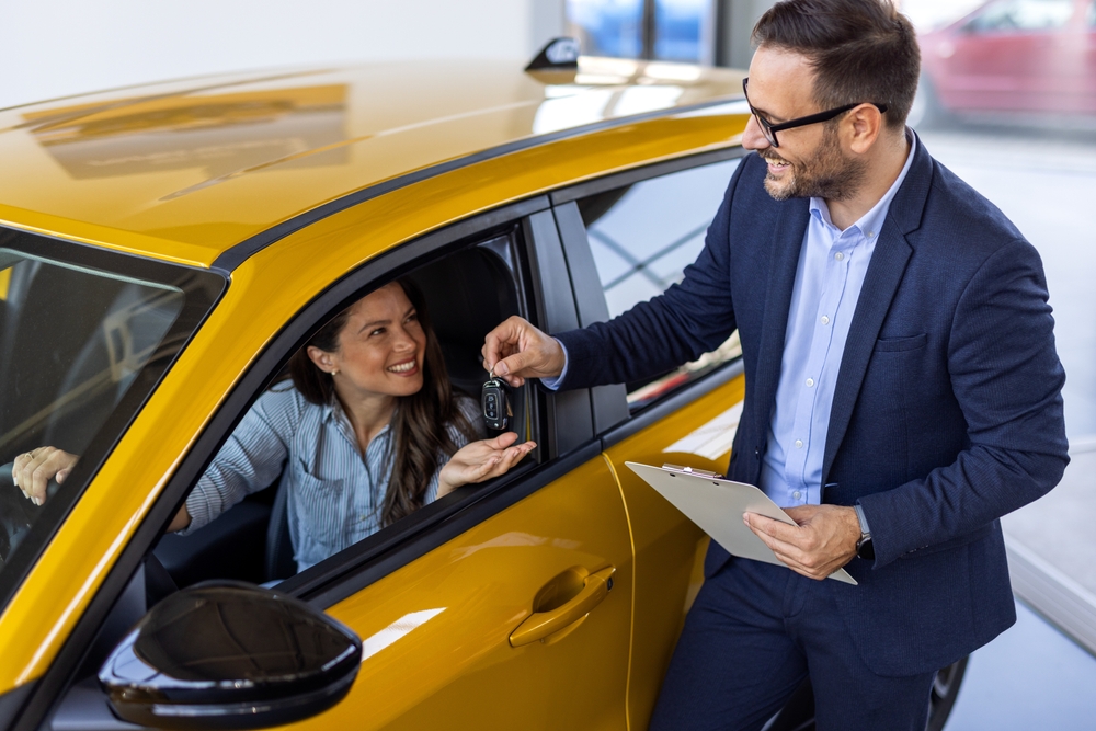 A woman behind the wheel of a yellow car smiles as she accepts the keys handed to her by a salesman at the car dealership.