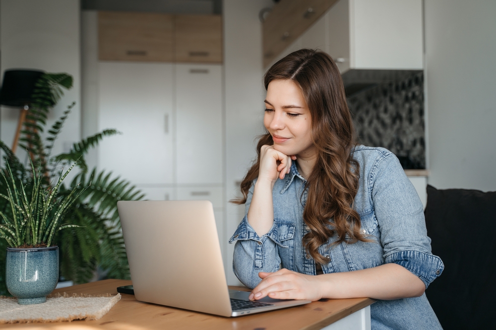 A woman with long brown hair and a blue shirt smiles and rests her chin on her hand as she checks her laptop.