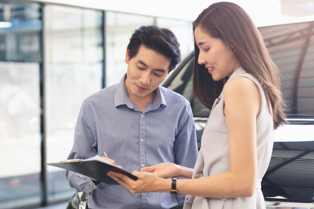 A woman with brown hair shows the leasing contract to a man in a blue shirt at a dealership showroom. 