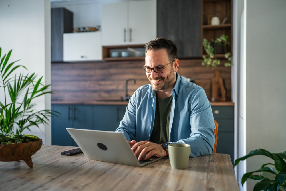 A smiling man wearing glasses sits at a kitchen table, an open laptop in front of him and a cup of coffee beside him.