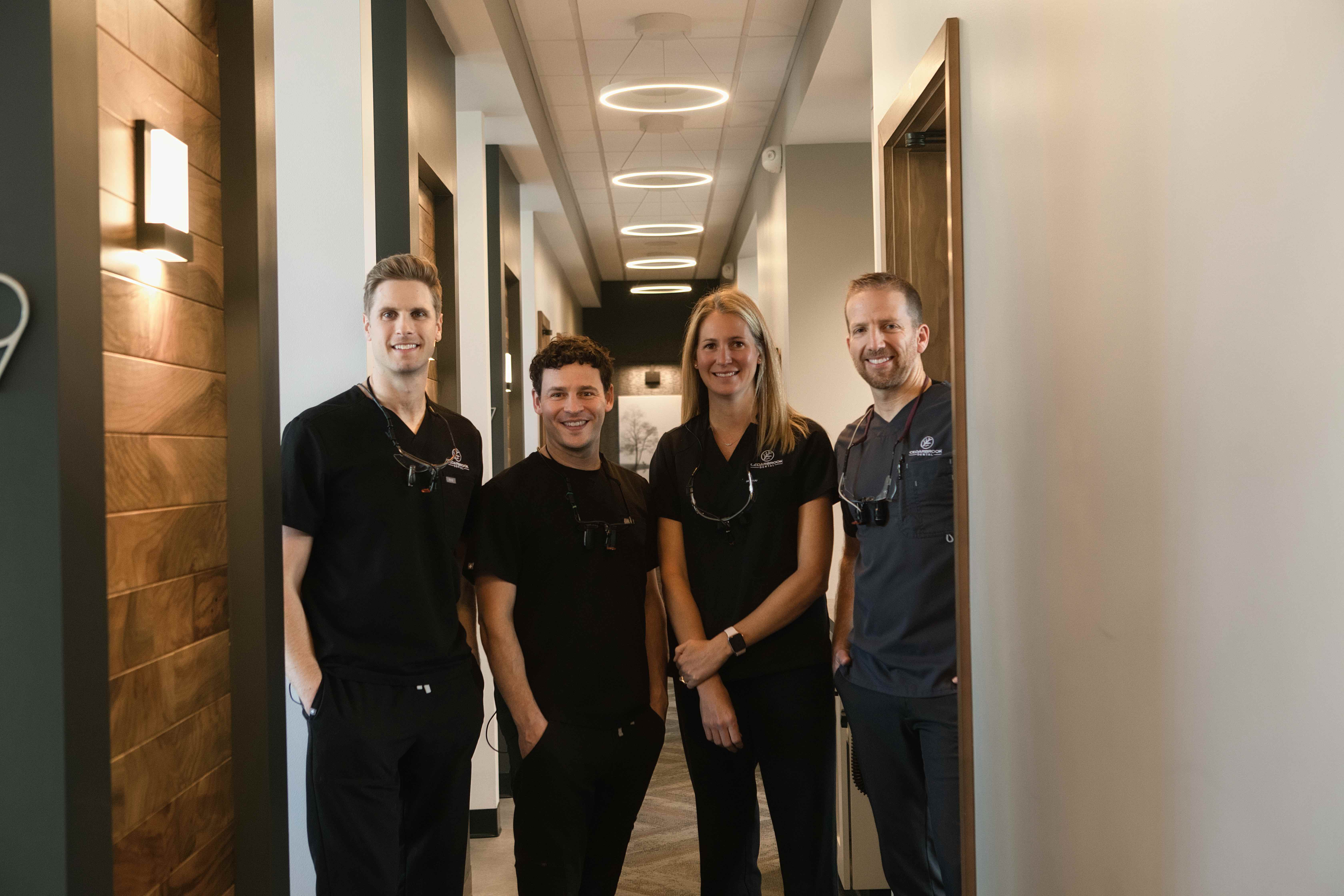 Four dental professionals in scrubs stand together and smile in a clinic hallway with modern lighting and decor.