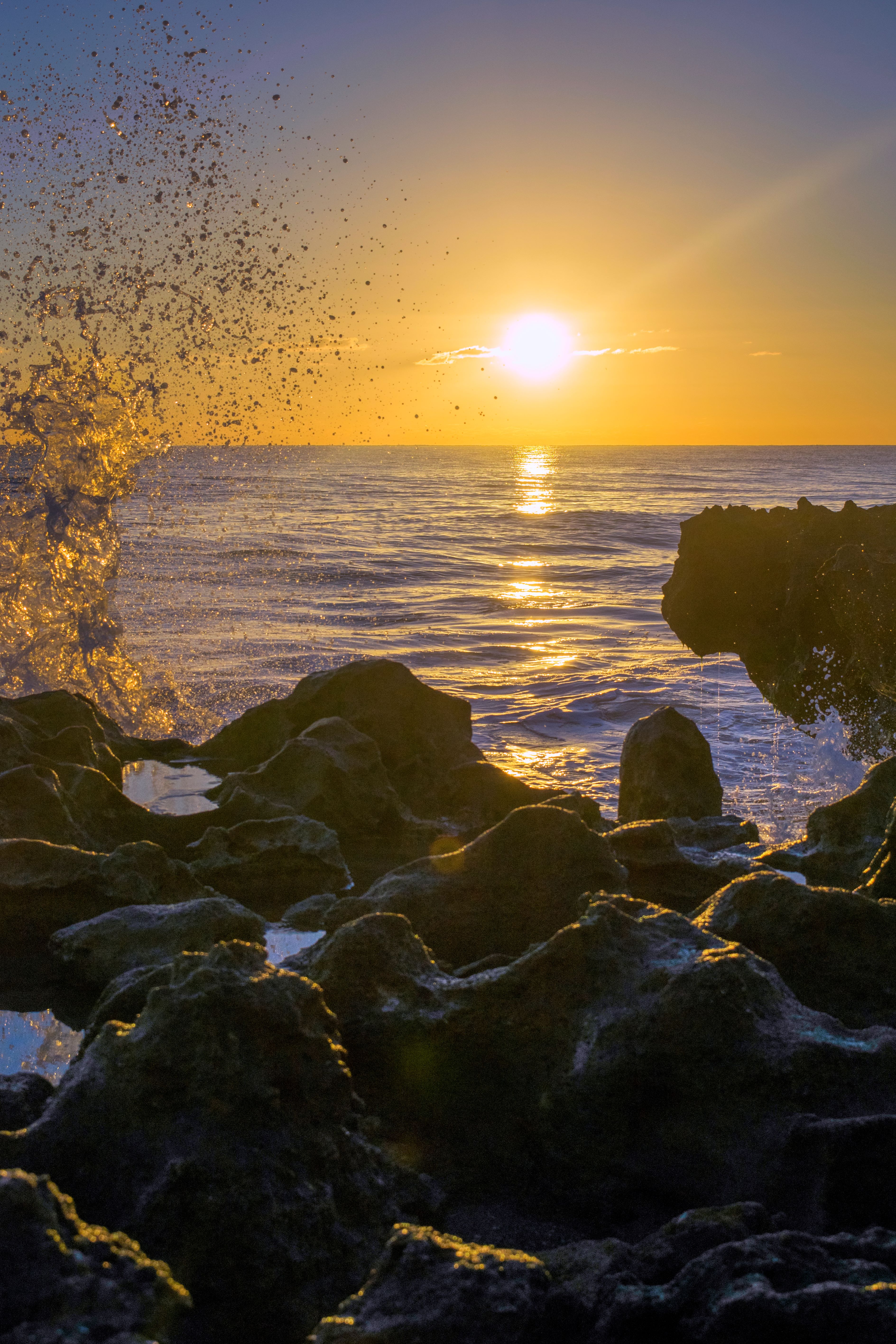 Photograph of a wave splashing above the coral rocks in front of the sunrise.