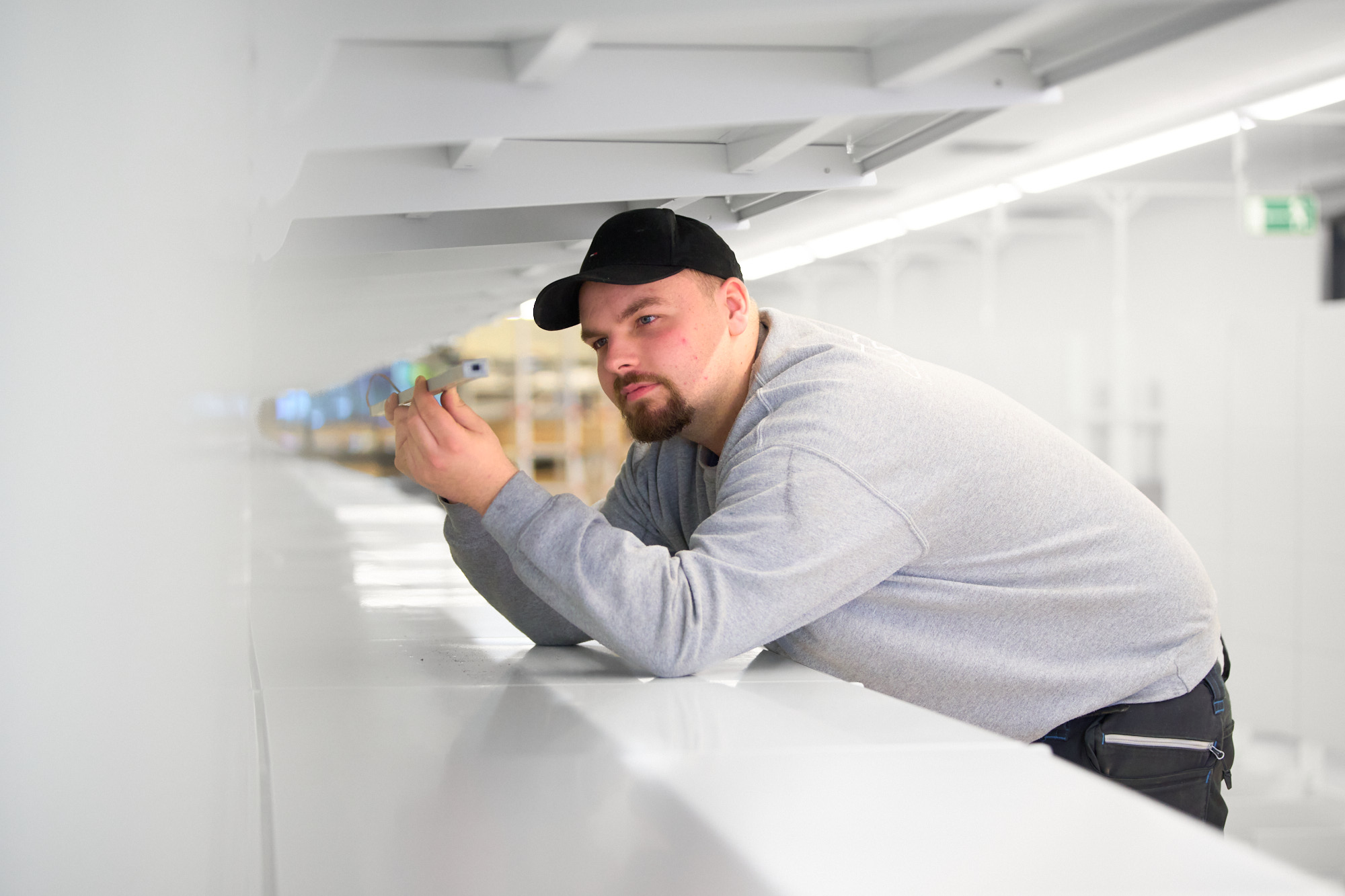 Electrician wearing a black cap and gray sweatshirt leaning over a white surface examining a small electronic device or component held in his hands in a brightly lit indoor space.