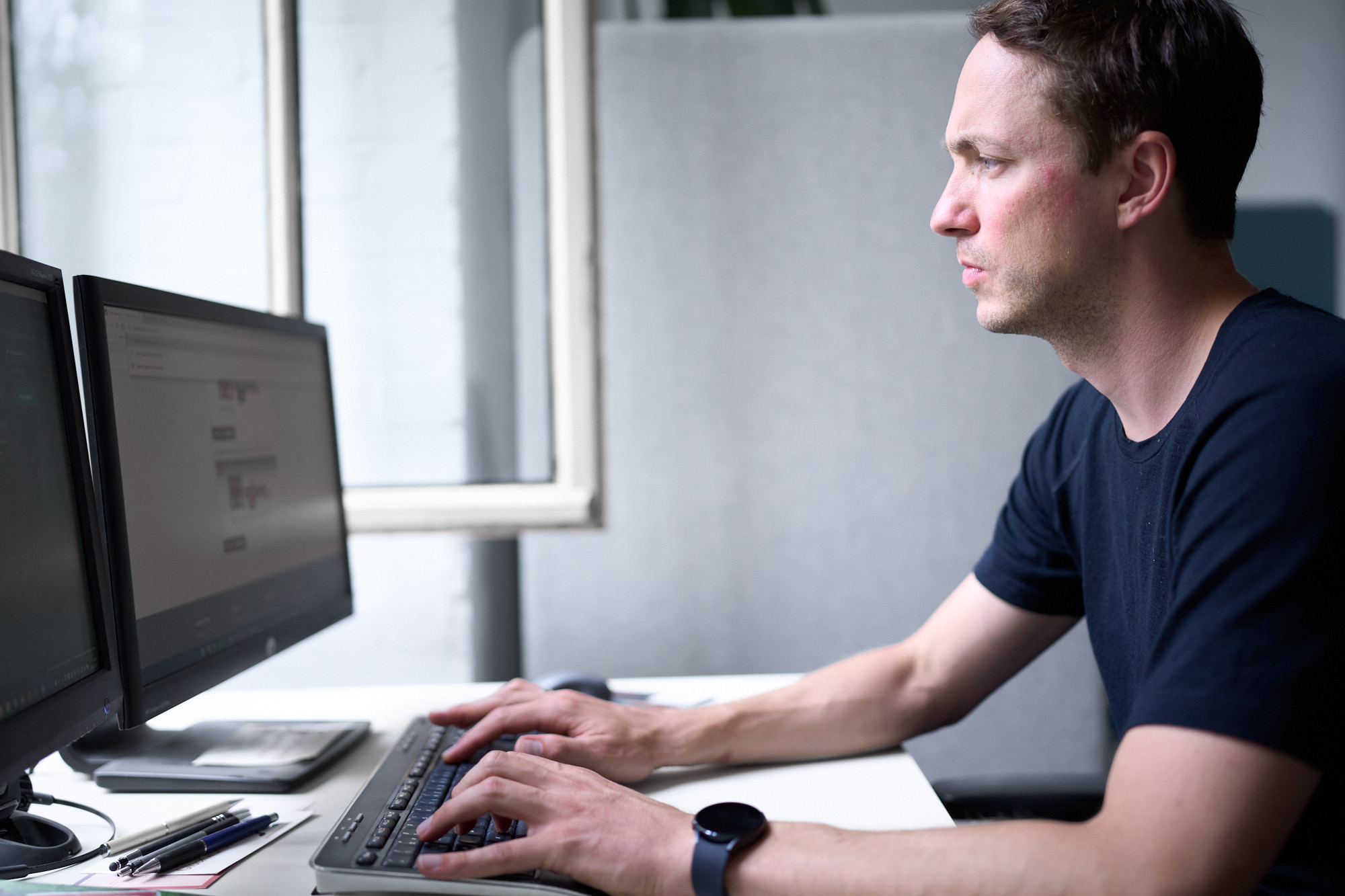 Man in black shirt sitting at a desk typing on a keyboard with two computer monitors in front of him, near a window.