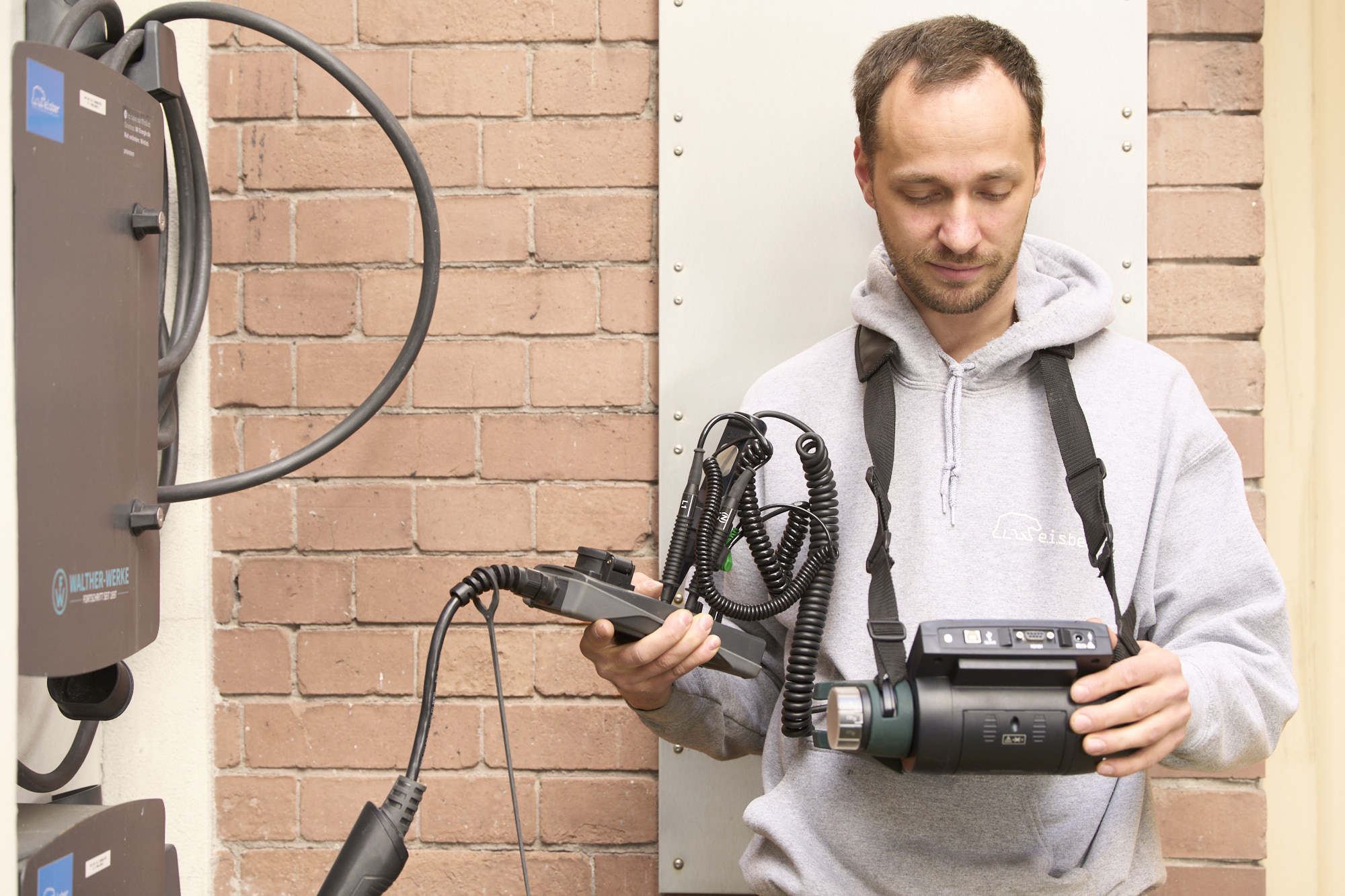 A man wearing a gray hoodie inspects a handheld device connected by coiled cables to an electric vehicle charging station mounted on a brick wall.