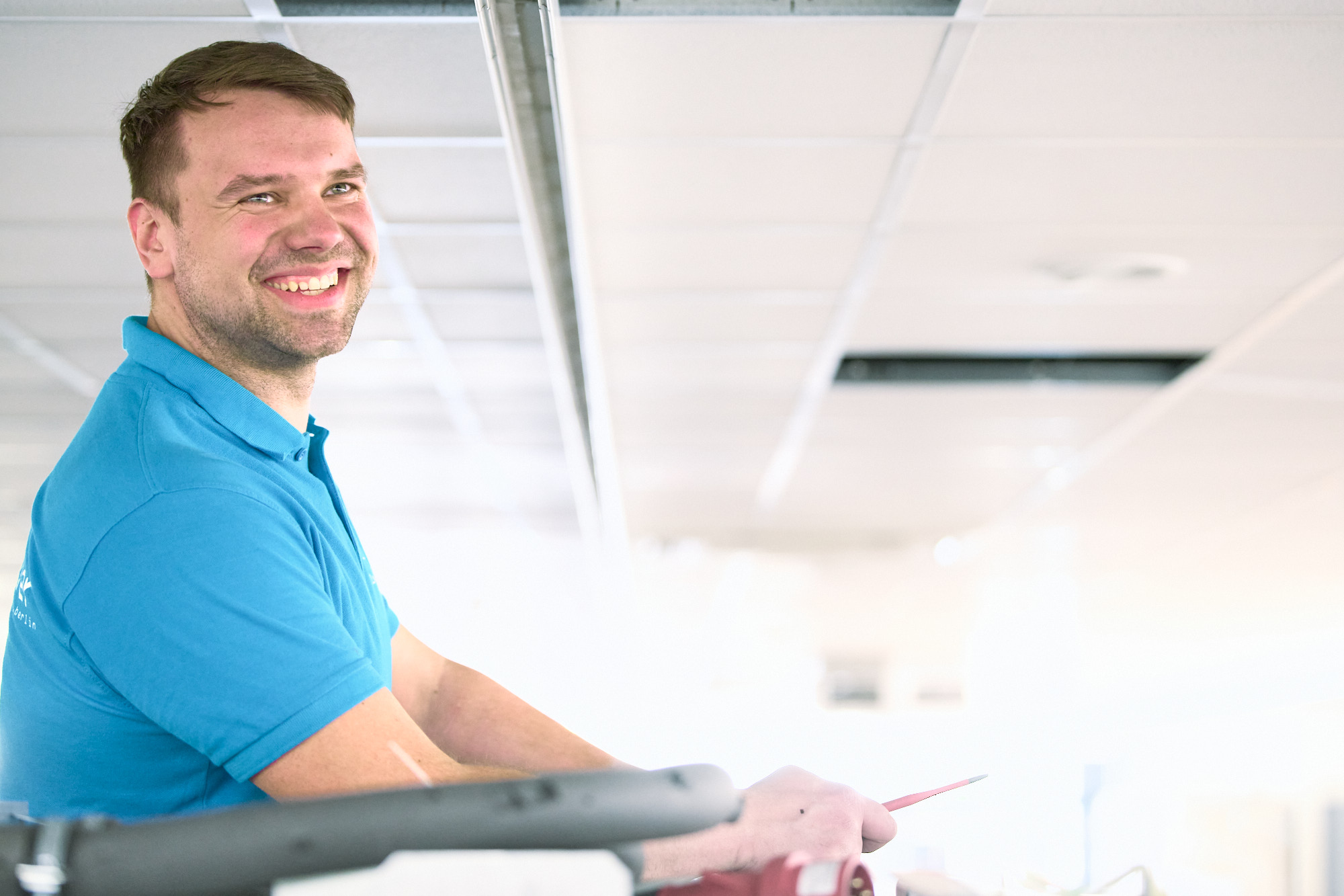 Smiling Electrician in a blue polo shirt standing in a bright indoor setting, holding a red tool or object in his right hand.
