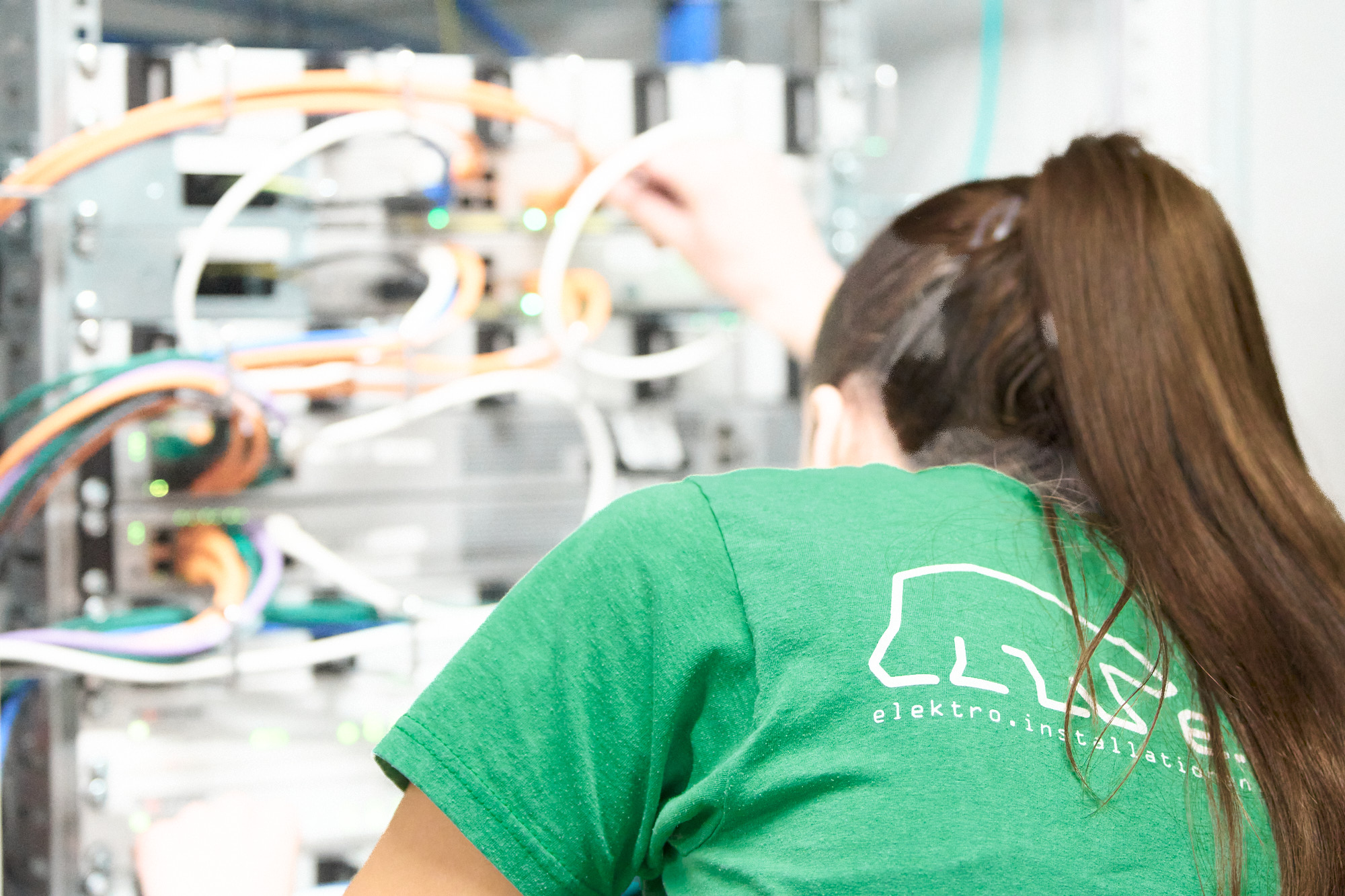 Woman with long brown hair tied in a ponytail wearing a green shirt with a white bear logo and text 'elektro.installation' on the back, working with orange, white, blue, and green cables connected to a server or network equipment panel.