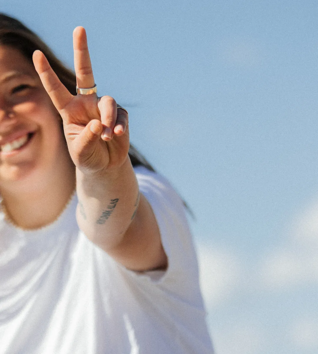 Smilende person i hvid t-shirt viser fredstegn mod himlen.