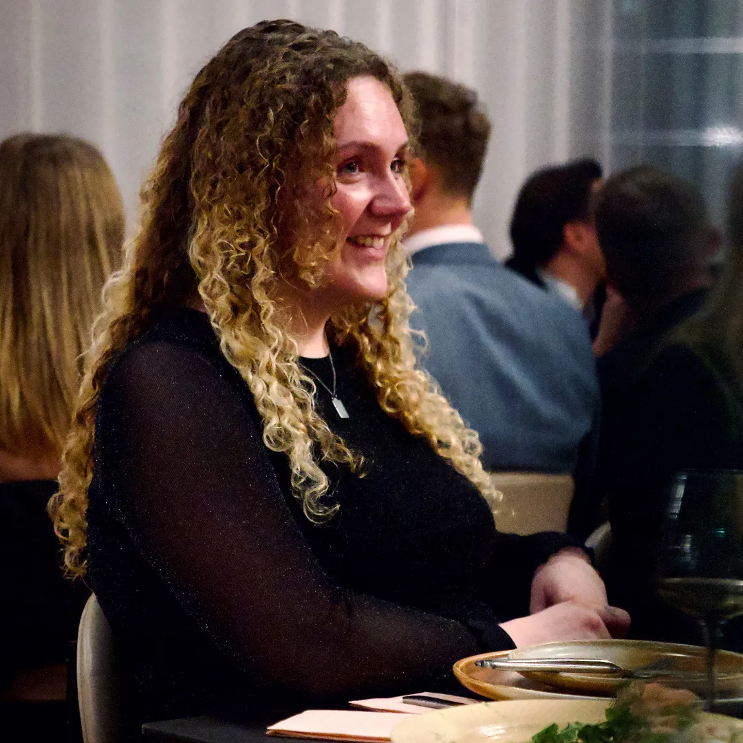 Kvinde med langt krøllet blondt hår smiler og sidder ved et bord med tallerkener og vinglas i et socialt arrangement.