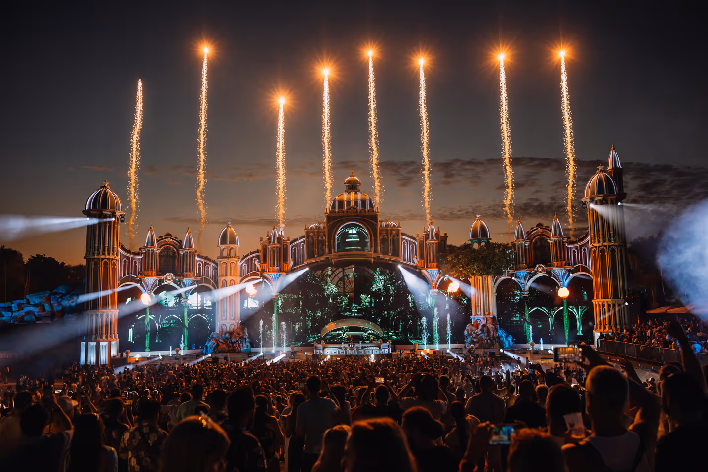 Large crowd watching a brightly lit, ornate stage with fireworks shooting into the night sky.