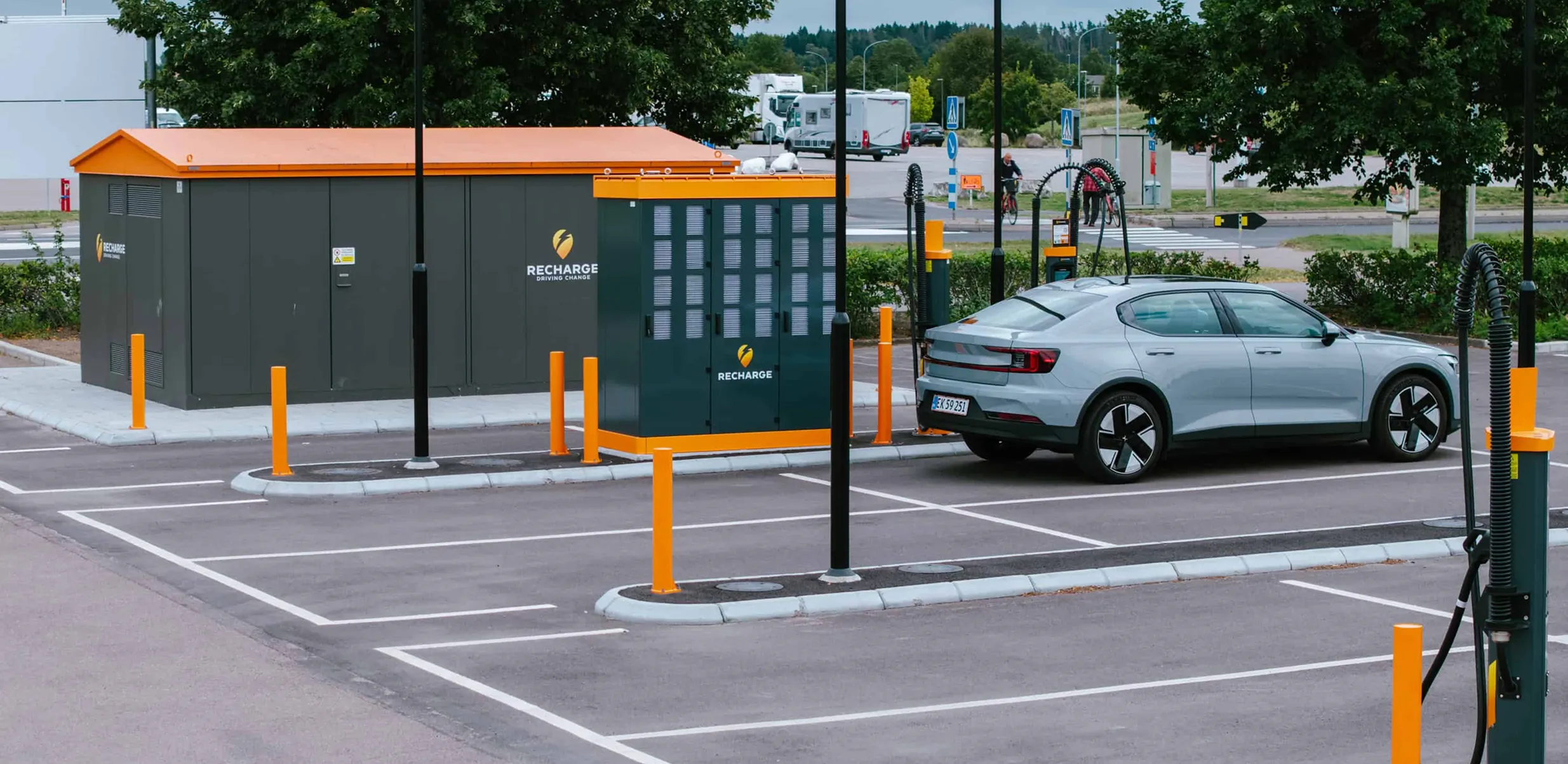 Silver electric car charging at a station with Recharge-branded equipment in a mostly empty parking lot.