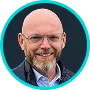 Smiling bald man with glasses and a beard wearing a dark jacket and blue shirt in a turquoise circular frame.