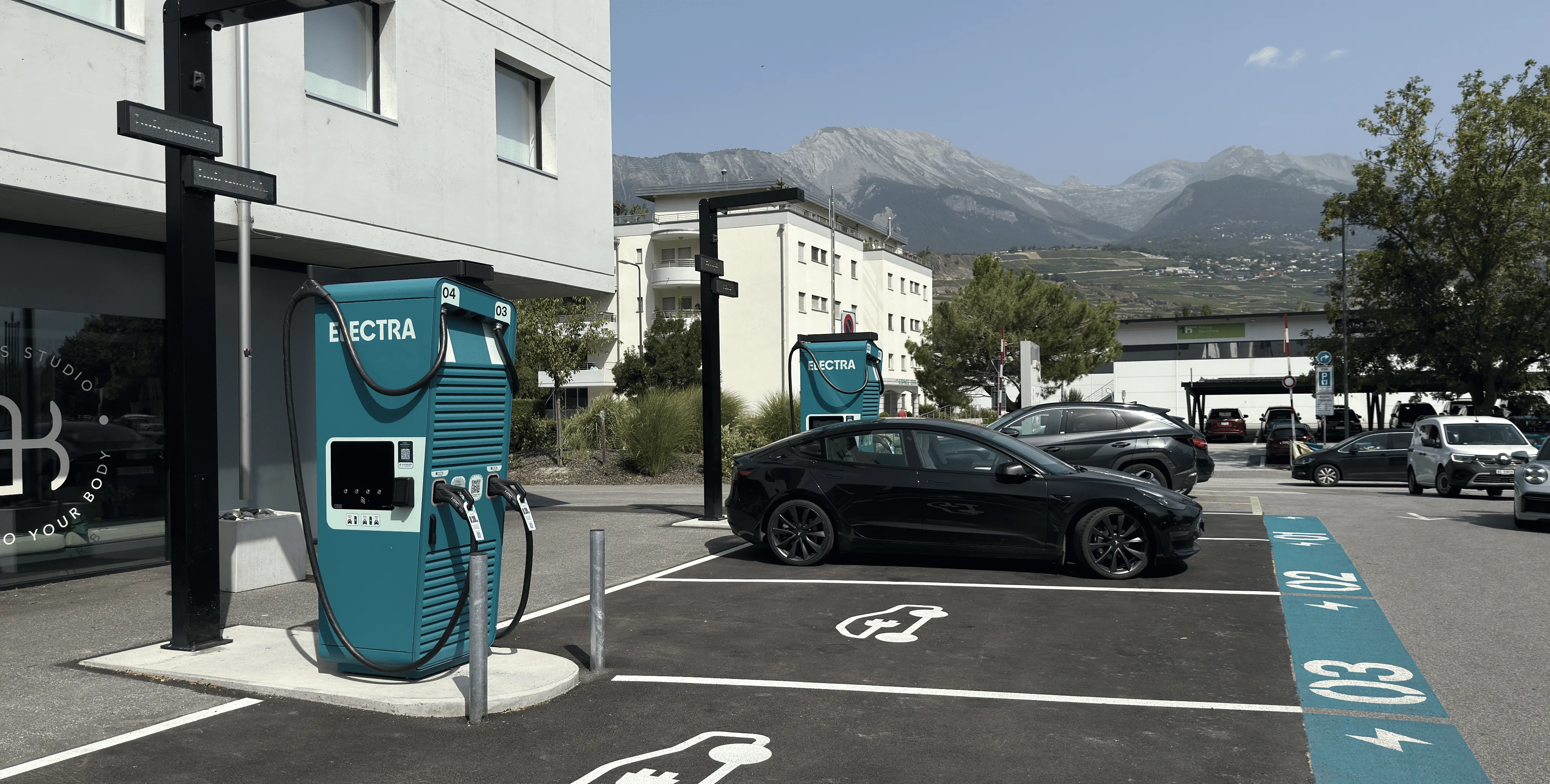 Electric vehicle charging station with two teal chargers labeled ELECTRA and a black electric car parked at one of the spaces with mountain scenery in the background.