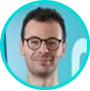 Smiling man with glasses and short brown hair in a turquoise circular frame.