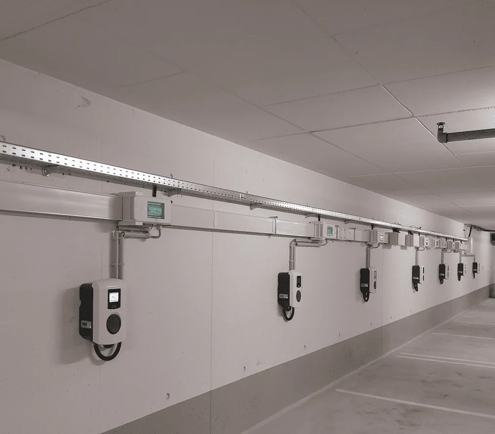 Indoor parking area wall with multiple electric vehicle charging stations evenly spaced and connected by overhead wiring.