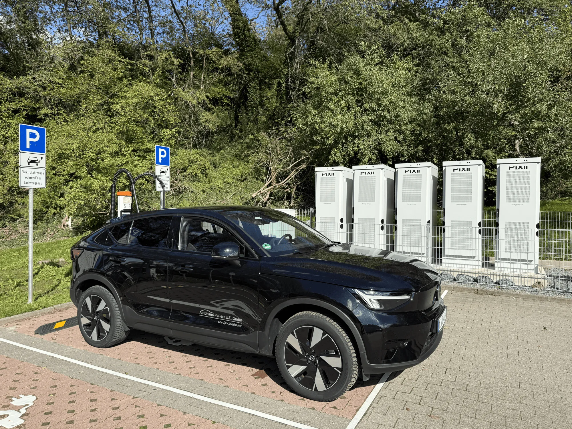 Black electric SUV parked at a charging station with multiple PXII energy storage units and parking signs in a wooded area.