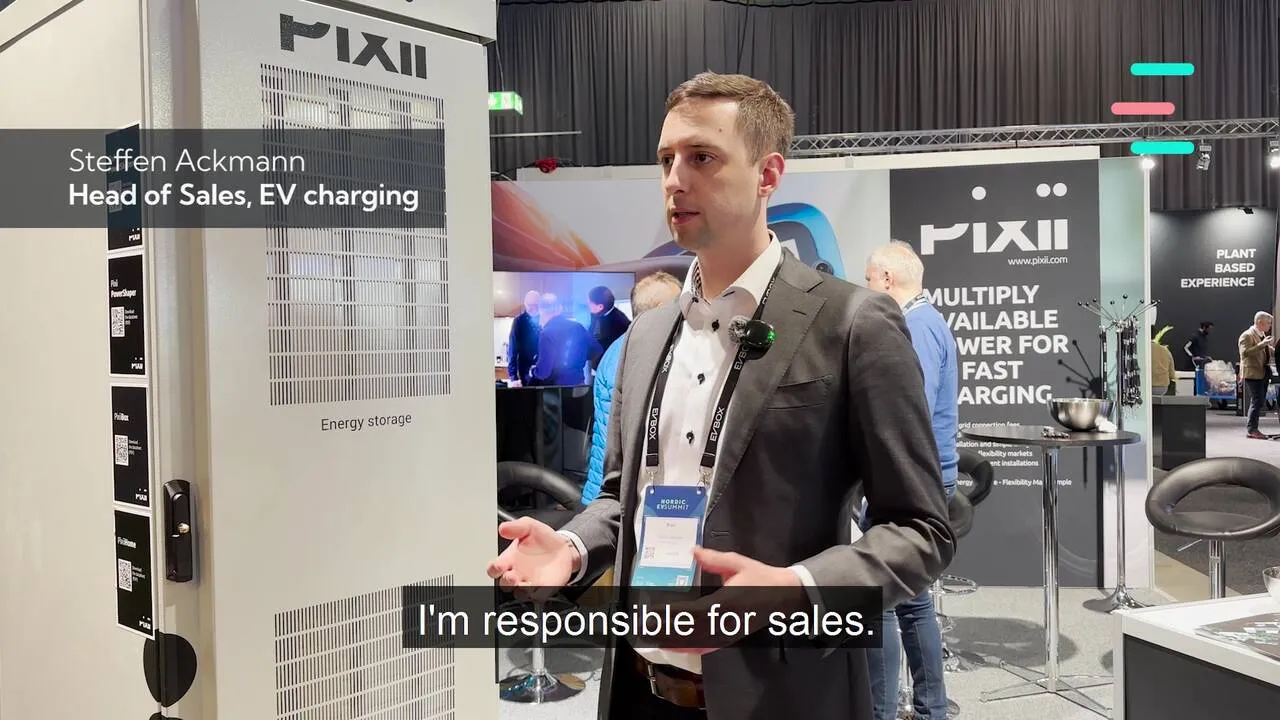 Man in a suit, wearing a conference badge, speaking at a booth with PIXII branding about EV charging sales.