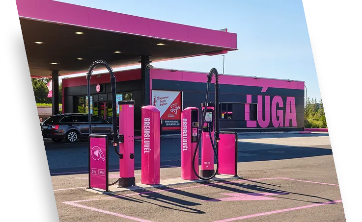 Bright pink electric vehicle charging station with multiple chargers under a matching pink canopy and a building in the background with large text 'LÚGA'.