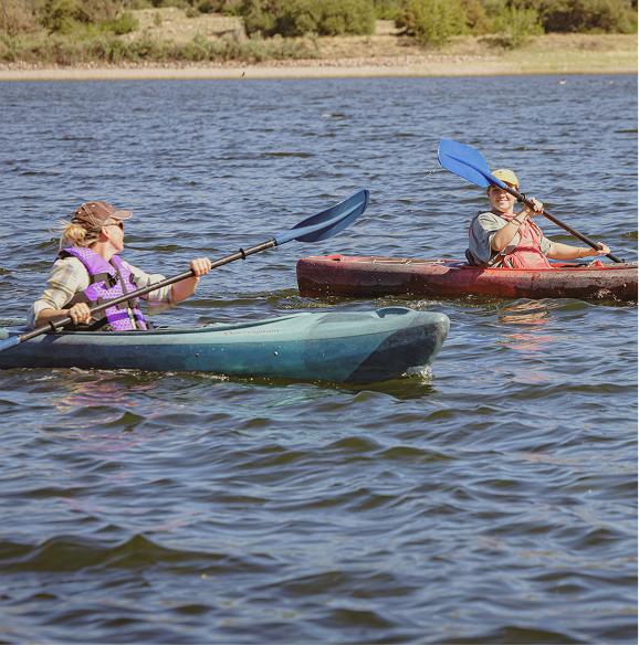 Two people kayaking on a calm lake wearing life jackets and hats under a sunny sky.