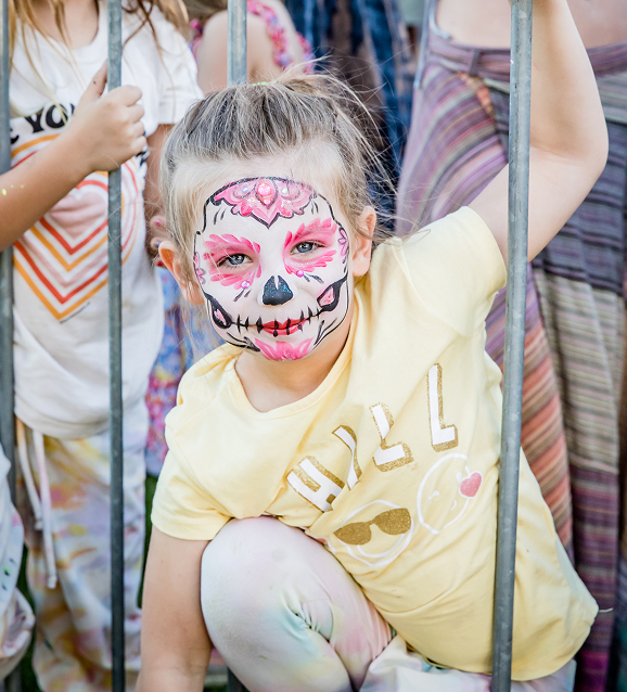 Child with pink and black skull face paint wearing a yellow shirt, holding metal bars.
