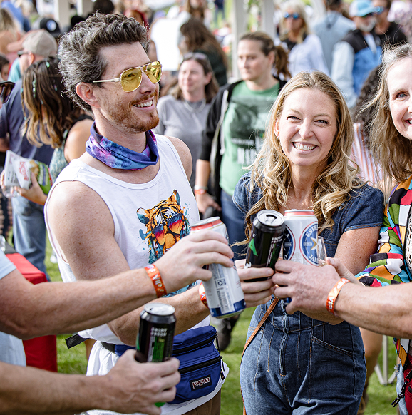 Group of people smiling and clinking canned drinks at an outdoor social event.