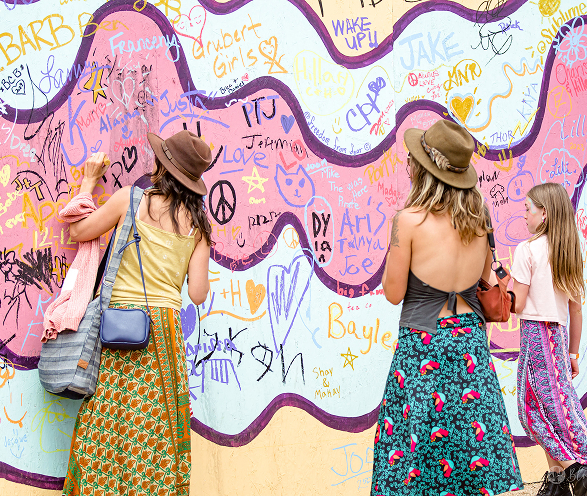Three females in colorful skirts and hats drawing and writing on a vibrant graffiti wall.