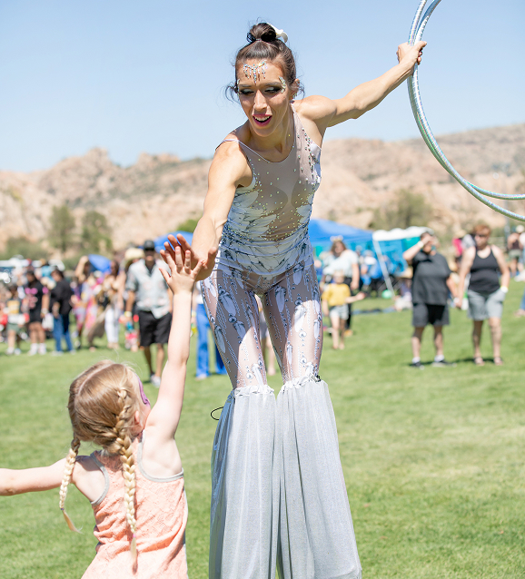 Woman on stilts in a sparkly costume high-fives a young girl with braided hair outdoors on grass.