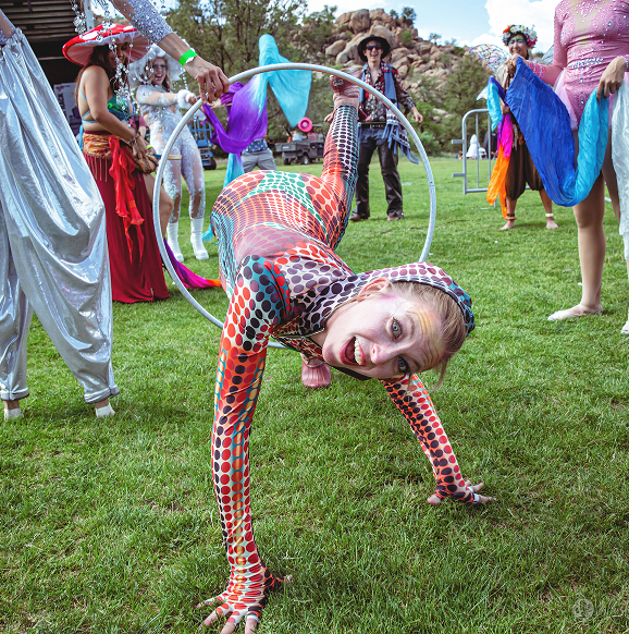 Person in colorful dotted costume crawling through a hula hoop on grass with costumed people around.