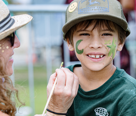 Boy smiling with green face paint being applied by a woman wearing a straw hat outdoors.