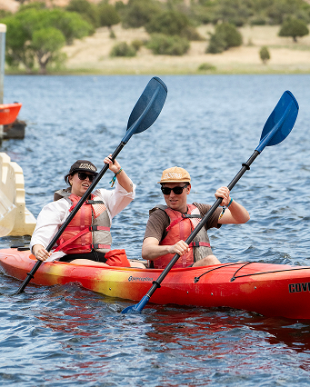 Two people kayaking on a calm lake wearing life jackets and hats under a sunny sky.