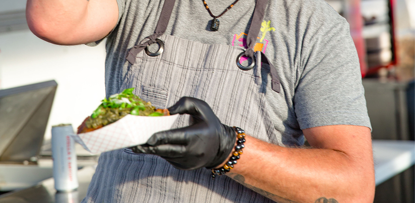 Smiling man in sunglasses and apron sprinkles green garnish on food in a white tray at a food stall.