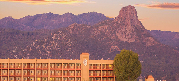 Building in front of forested mountain with a tall rocky peak during sunset.