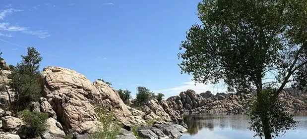 Rocky terrain beside a calm lake with green trees under a blue sky with light clouds.
