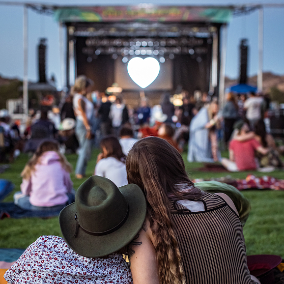 Couple sitting on grass at outdoor concert with illuminated heart shape on stage in the background.