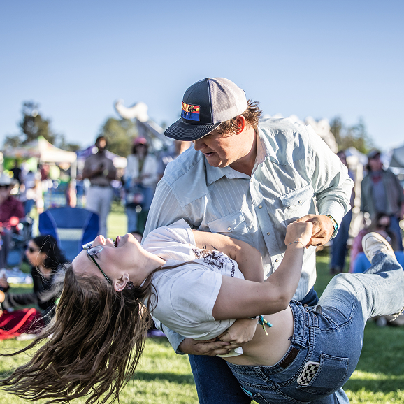 Man in cap dips laughing woman with glasses during outdoor event on grassy field.