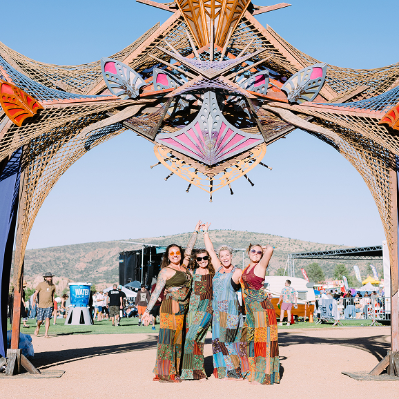 Four women in colorful patchwork dresses posing and smiling under a large wooden butterfly arch at a festival.