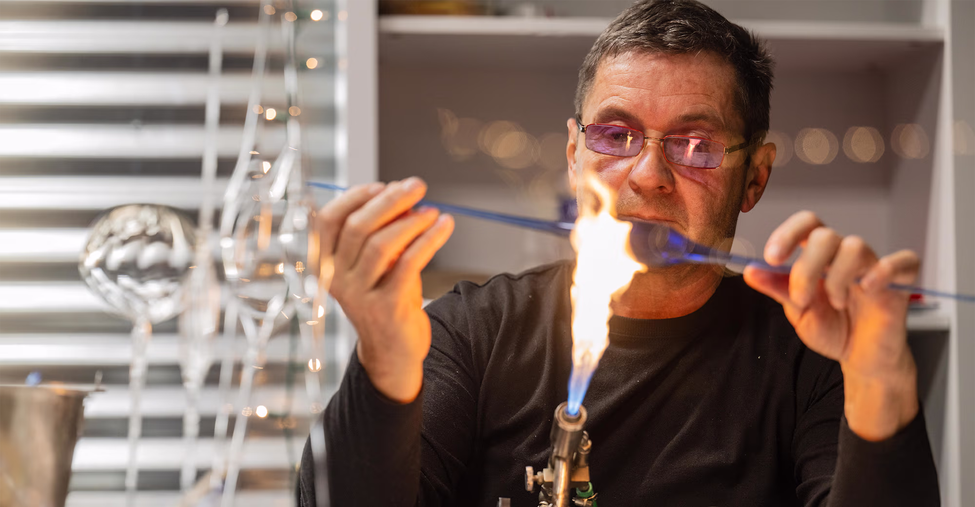 Man wearing glasses shaping glass tubing with a flame torch in a workshop.