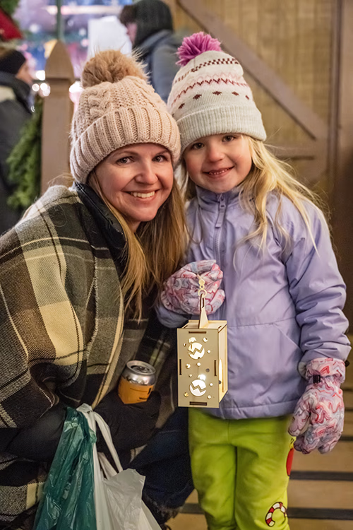 Smiling woman and young girl wearing winter clothes and knit hats, with the girl holding a decorative lantern.
