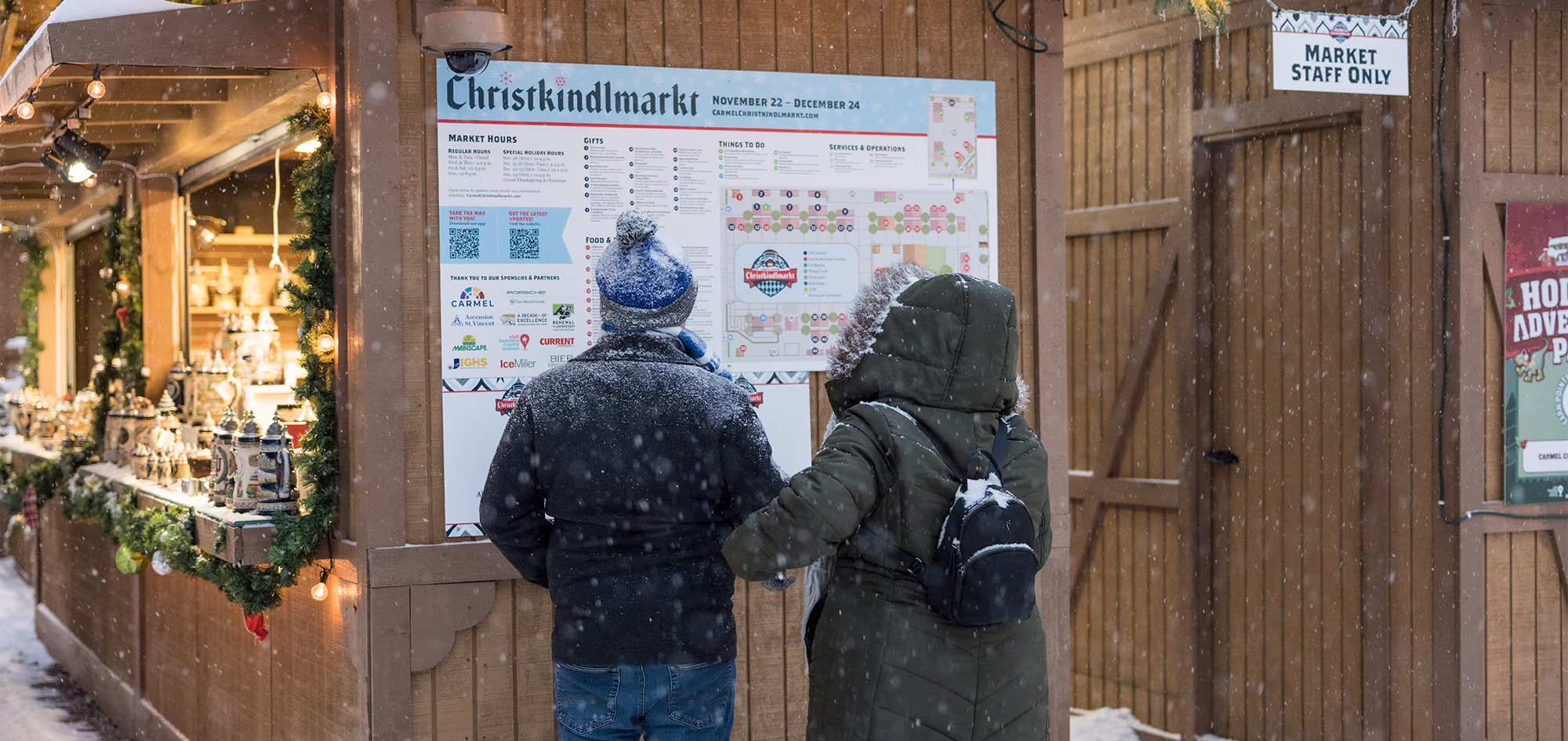Two visitors read a Christkindlmarkt map beside a snow-covered, warmly lit market stall.