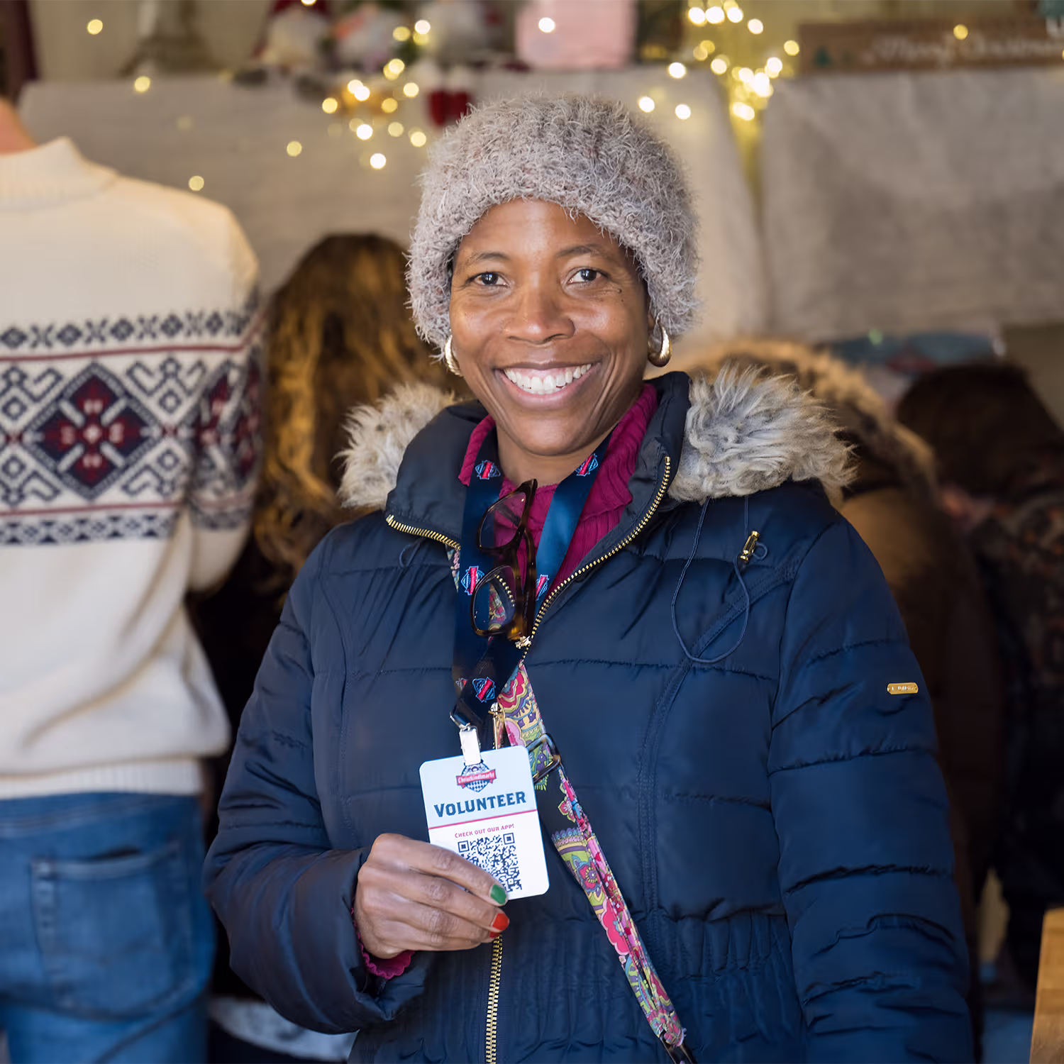 A Christkindlmarkt volunteer smiles while holding her badge inside a busy booth.