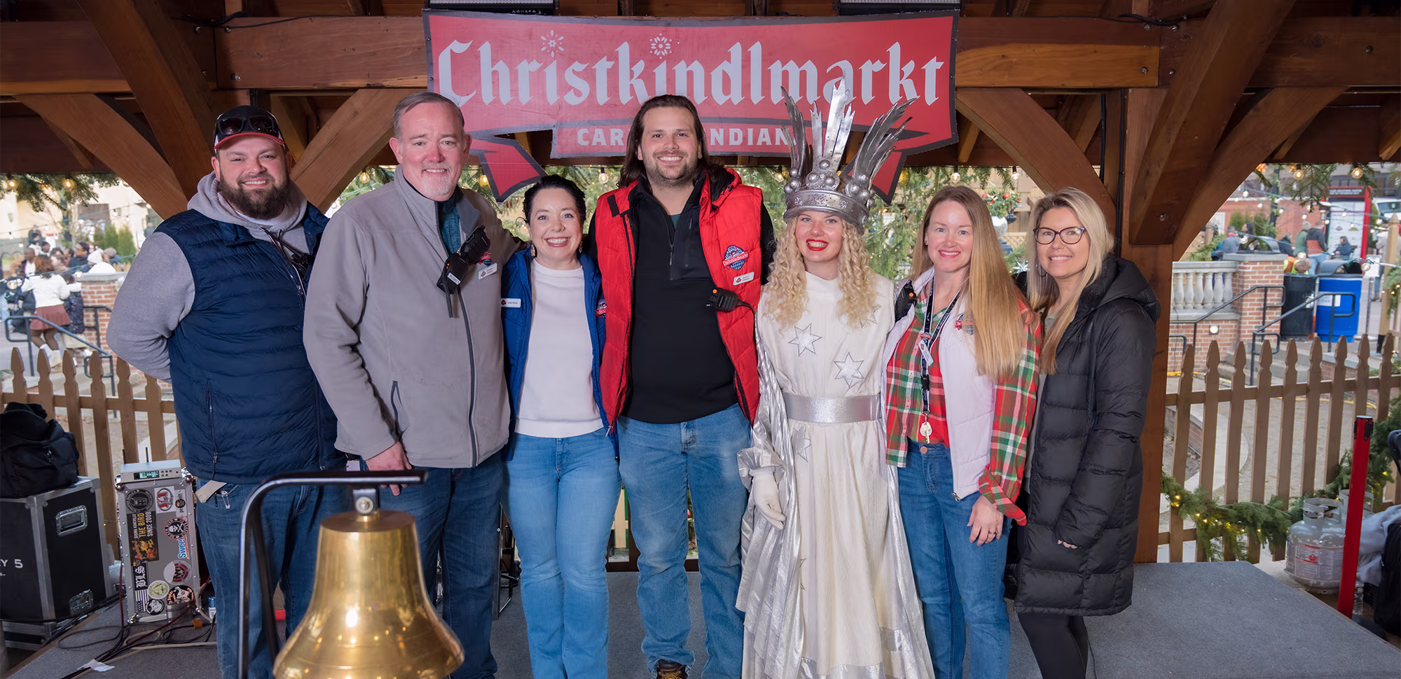 A group poses on the Christkindlmarkt stage with a costumed holiday character and a brass bell.