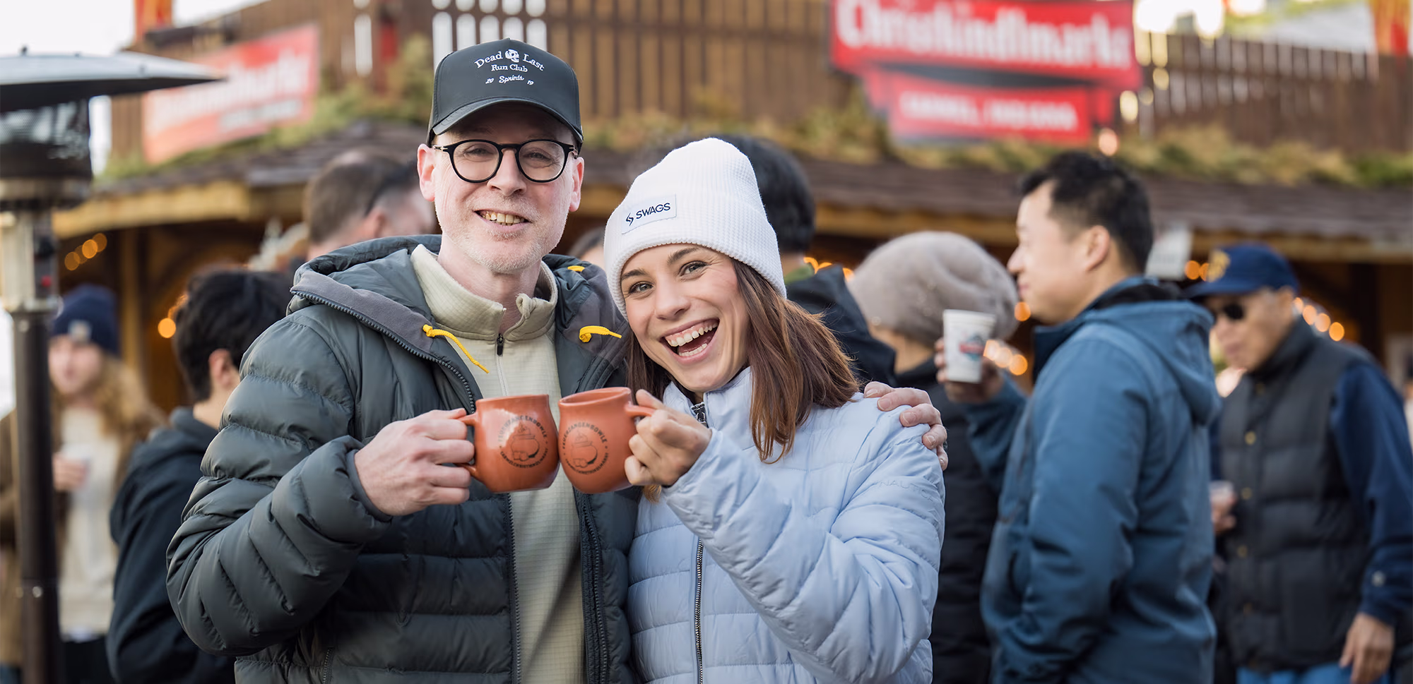 Two visitors toast with mugs at a busy outdoor Christkindlmarkt.