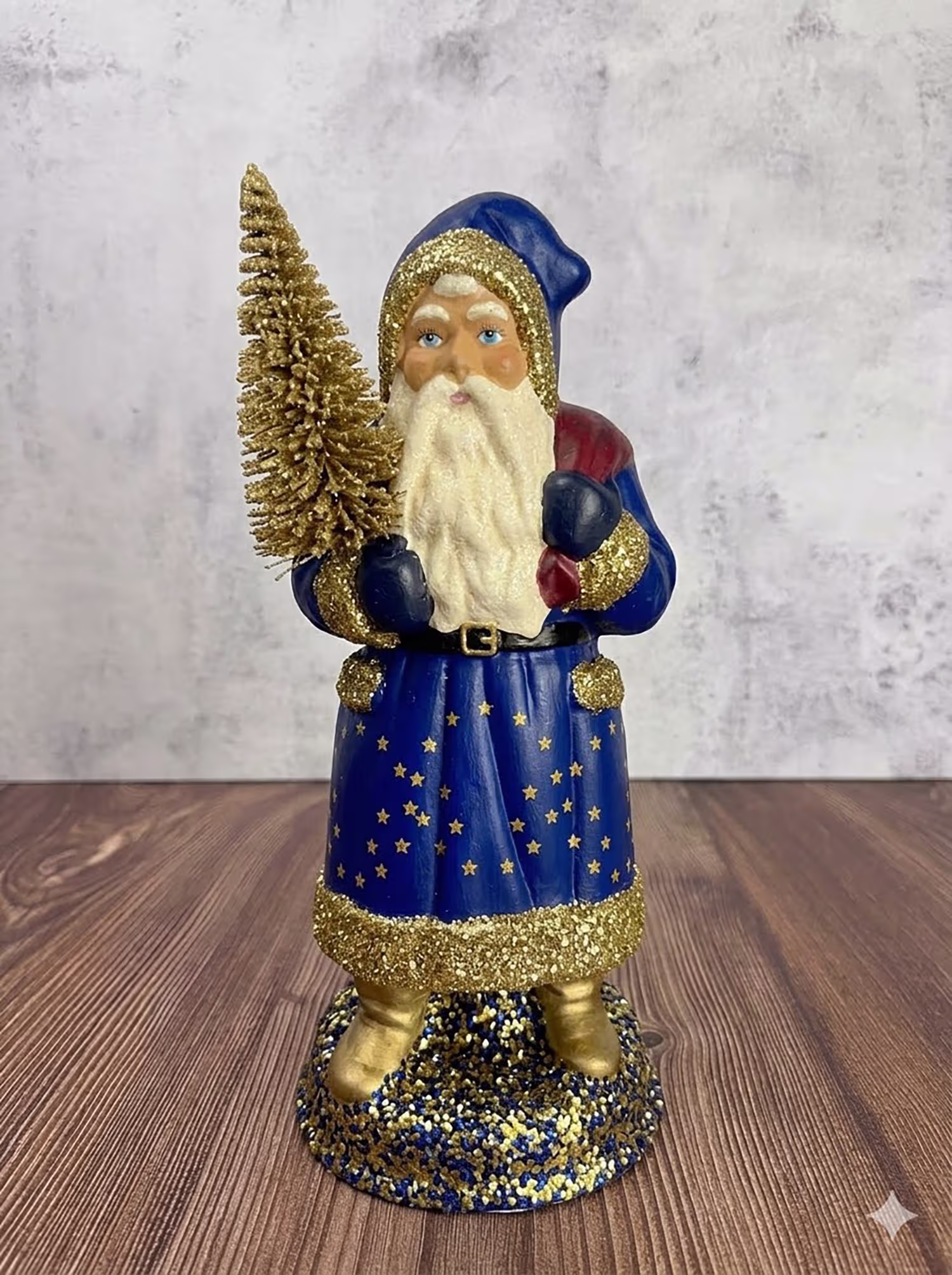 Figurine of Santa Claus wearing a blue robe decorated with gold stars and glitter trim, holding a glittery gold Christmas tree in one hand, standing on a round glittery base on a wooden surface with a gray textured background.