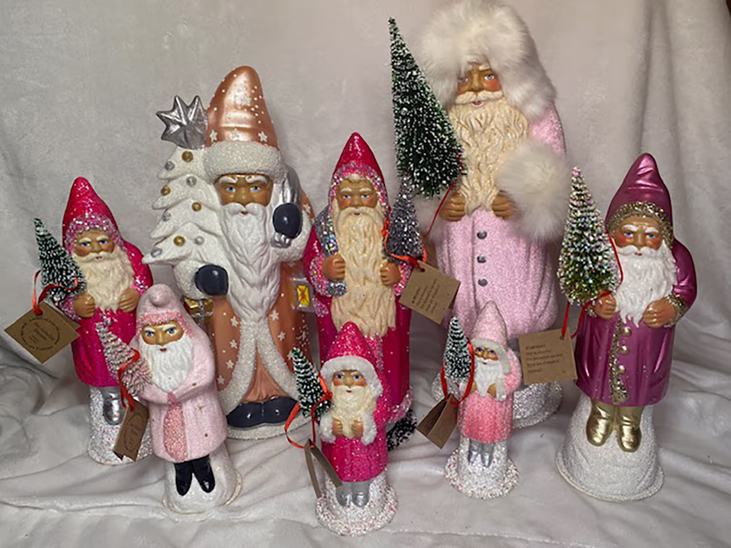 Group of eight decorative Santa Claus figures in various pink and purple outfits holding small snow-covered Christmas trees, arranged on a white fabric background.